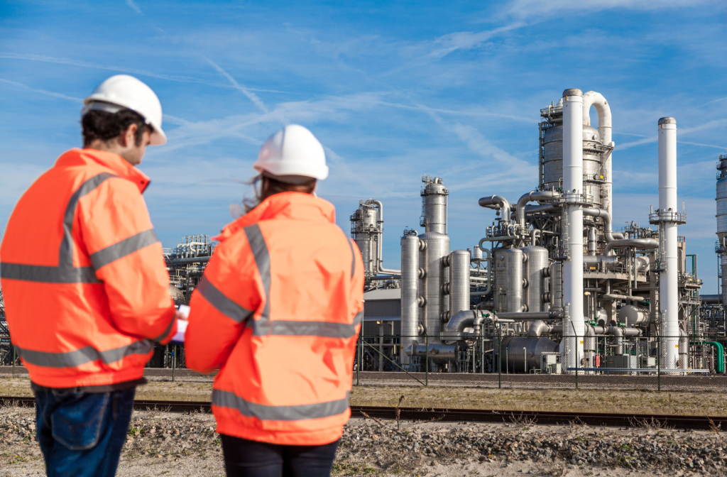 Profissionais com capacete e jaqueta de alta visibilidade diante de uma planta industrial.